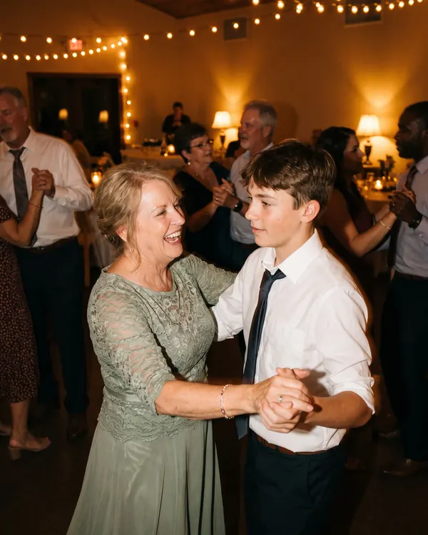 A grandmother in a lace dress laughs as she dances with her teenage grandson.