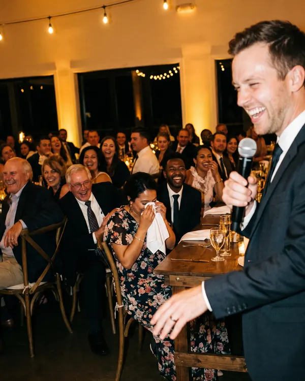 The best man mid-toast with a microphone while the long table laughs, lit by a camera flash.