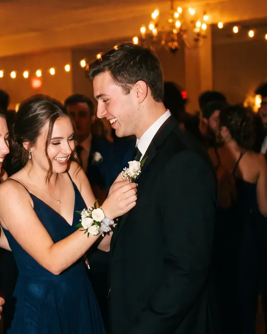 A young woman in a navy gown pins a white boutonniere on her date’s lapel on a crowded dance floor.